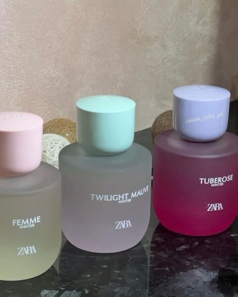 Trzy okrągłe flakony perfum Zara w matowym, szronionym szkle, w zimowej edycji "Winter", stojące na ciemnej powierzchni. Zapachy to "Femme Winter" (delikatny żółty płyn i jasnoróżowy korek), "Twilight Mauve Winter" (płyn w odcieniu przydymionego różu i miętowy korek) oraz "Tuberose Winter" (intensywnie różowy płyn i lawendowy korek). Tło jest rozmyte, z widocznymi dekoracyjnymi kulami ze sznurka.