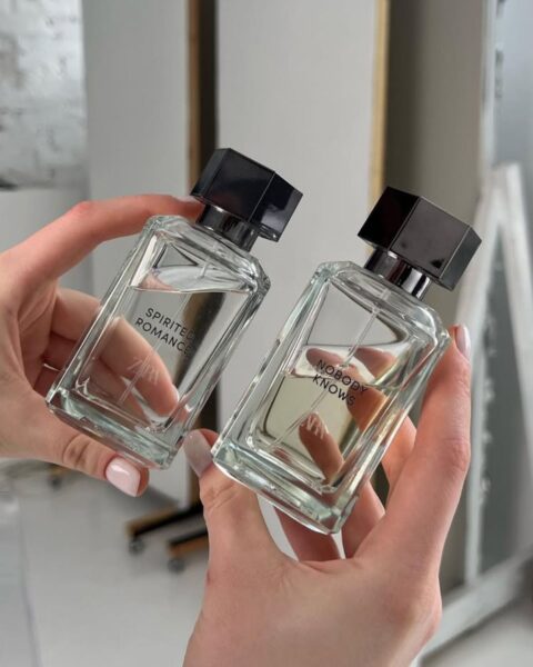 Dwie dłonie o jasnym manicure trzymają dwa prostokątne flakony perfum Zara w minimalistycznym, przezroczystym szkle z ciemnymi, geometrycznymi korkami. Na butelkach widoczne są napisy "Spirited Romance" i "Nobody Knows". Tło jest jasne, nowoczesne i minimalistyczne, z elementami białych mebli i lustrzanych powierzchni.