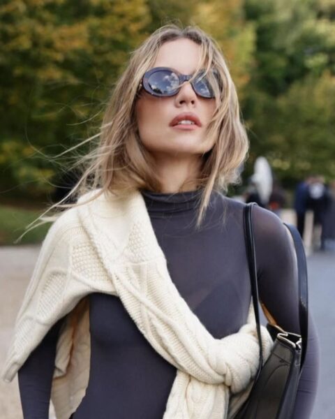 Elegancka kobieta w szarym golfie i modnych, owalnych okularach przeciwsłonecznych. Na ramionach ma luźno narzucony kremowy sweter o warkoczowym splocie. Włosy rozwiewa jej delikatny wiatr, a stylizacja jest idealna na wczesną jesień. Ten look doskonale komponowałby się z ciepłymi i otulającymi perfumami pachnącymi babim latem.