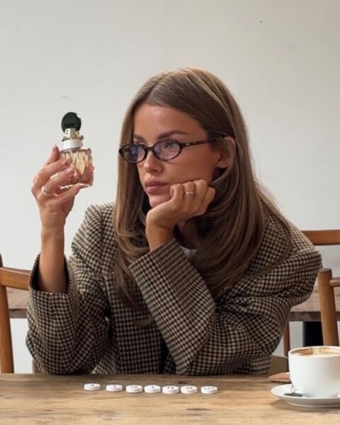 Elegancka kobieta w okularach i marynarce w pepitkę, siedząca przy drewnianym stole. Trzyma w dłoni flakon perfum i zamyślona patrzy w bok. Obok niej stoi filiżanka kawy.