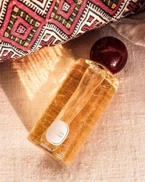 Luksusowy flakon perfum Benjoin Bohème Diptyque w ciepłym, złocistym świetle, leżący na beżowym lnianym tle. Butelka ma unikalną, spękaną teksturę szkła i jest zwieńczona bordowym korkiem. Obok leży fragment tkaniny z tradycyjnym, barwnym wzorem w odcieniach czerwieni i żółci. Idealna kompozycja wizualna i zapachowa dla kobiet ceniących orientalne nuty, w tym perfumy z nutą cynamonu.