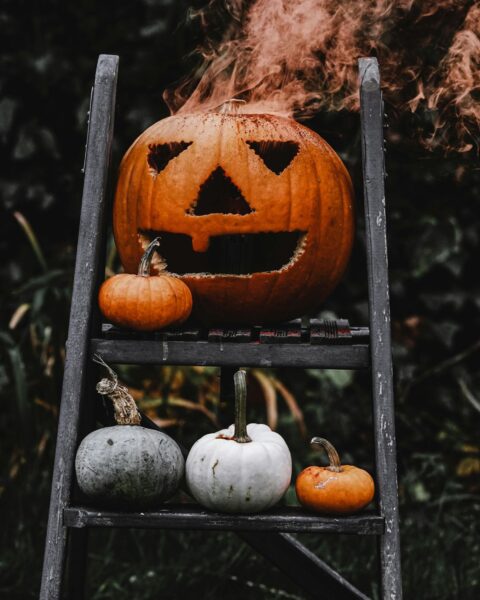Mroczny kadr pionowy. Duża pomarańczowa dynia z wyciętą straszną twarzą (jack-o'-lantern) stoi na drewnianej drabinie ogrodowej i wydobywa się z niej rdzawoczerwony dym. Pod spodem, na niższym szczeblu, leżą mniejsze dynie: szara, biała i pomarańczowa, tworząc klimatyczną dekorację na Halloween.
