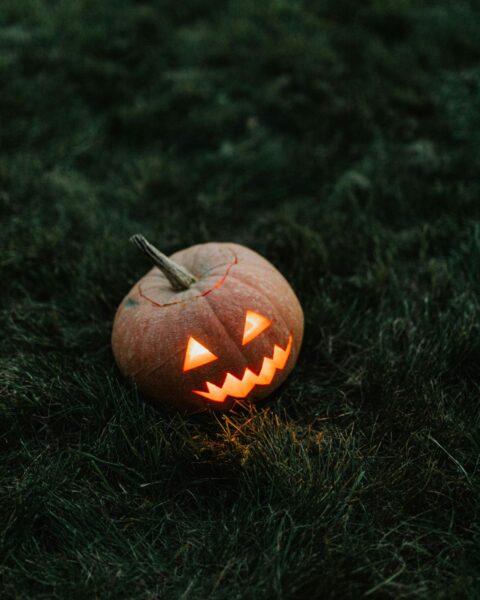 Mała, rzeźbiona dynia (jack-o'-lantern) z płonącym, pomarańczowym wnętrzem, leżąca w kadrze centralnym na ciemnej, soczyście zielonej trawie. Mroczny, minimalistyczny nastrój Halloween.