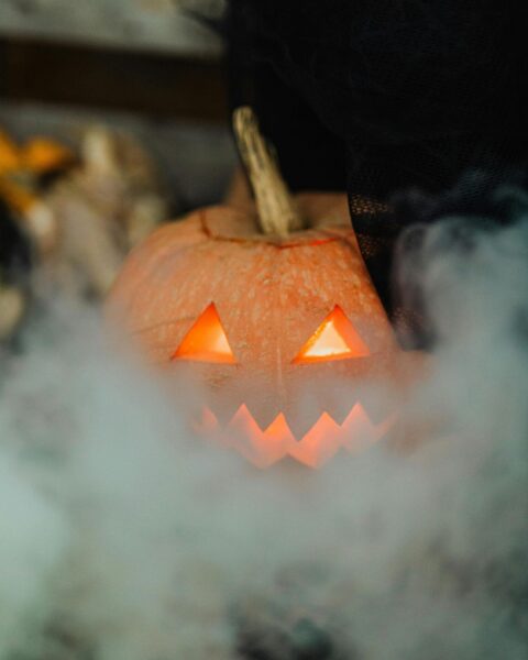 Zbliżenie na małą, pomarańczową dynię z wyciętymi w kształcie trójkątów oczami i zębami (jack-o'-lantern), która jest otoczona gęstą, białą mgłą lub dymem. Ciepłe światło z wnętrza dyni przebija się przez zasłonę dymu, tworząc tajemniczy, halloweenowy nastrój.