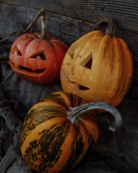 Martwa natura na Halloween: Trzy dynie w różnych odcieniach pomarańczu i żółci, ustawione na szarym, workowym materiale. Dwie z nich są wydrążone z wyciętymi przerażającymi twarzami (jack-o'-lantern), a trzecia, z tygrysim wzorem, leży na pierwszym planie. Klimatyczne dekoracje w tle postarzałego drewna.