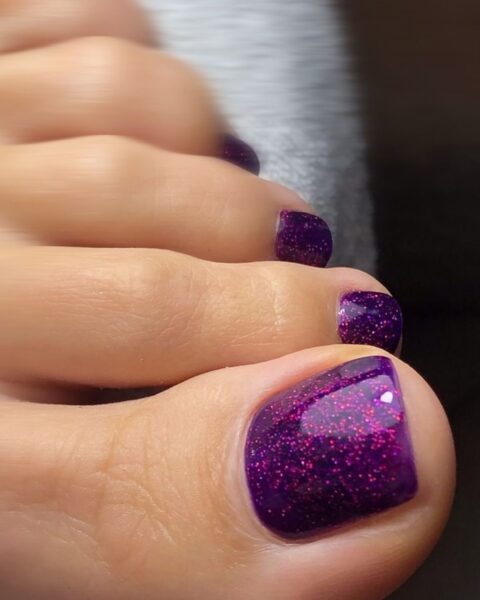 Pedicure hybrydowy w głębokim, intensywnym odcieniu śliwkowego fioletu, wzbogacony mnóstwem różowego i fioletowego brokatu. Paznokcie u stóp są lśniące i idealnie wykończone. Zdjęcie wykonane w dużym zbliżeniu na palce, eksponując trójwymiarowy, migoczący efekt brokatu. Elegancki i przyciągający wzrok pedicure wieczorowy.