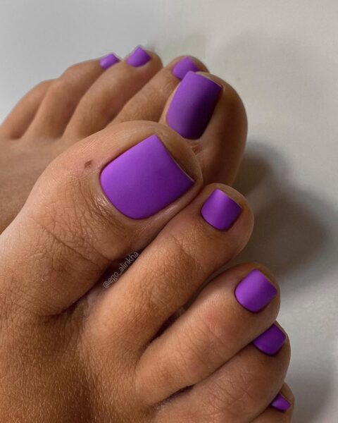 Modny matowy pedicure w intensywnym, soczystym fiolecie (odcień fuksji lub lawendy). Paznokcie u stóp są starannie wypiłowane do kształtu kwadratowego i pokryte matowym lakierem hybrydowym. Wykończenie jest idealnie gładkie i aksamitne, podkreślające wyrazistość koloru. Nowoczesny i odważny design paznokci.