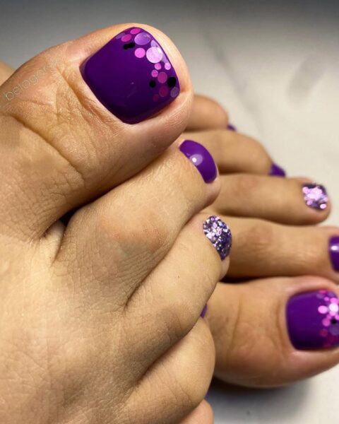 Fioletowy pedicure hybrydowy w intensywnym, nasyconym odcieniu fioletu na zadbanych stopach. Paznokieć dużego palca ozdobiony jest modnym wzorem z błyszczących cekinów/konfetti w odcieniach fioletu, różu i czerni. Mały palec również ma brokatowe wykończenie. Nowoczesny i odważny design paznokci. Ujęcie z bliska, skupione na precyzyjnym wykonaniu.