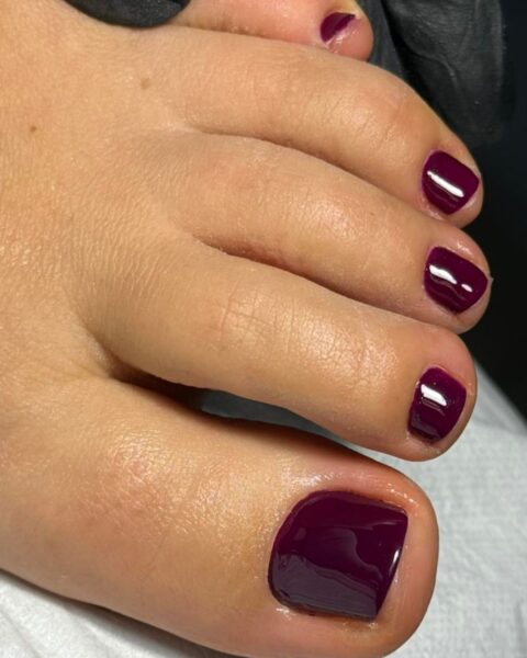 Perfekcyjny pedicure hybrydowy w głębokim, lśniącym odcieniu ciemnej śliwki/burgundu. Paznokcie u stóp są starannie opracowane, skrócone do idealnego, równego kształtu i pokryte błyszczącym lakierem z efektem lustrzanego połysku. Klasyczny i elegancki kolor idealny na jesień lub jako wyrafinowany element stylizacji.