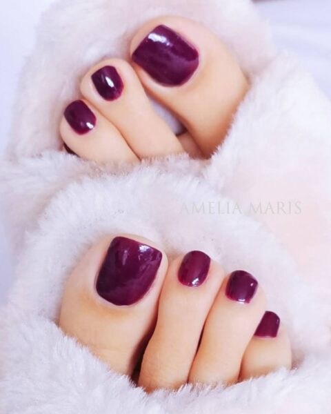 Zimowy pedicure w bogatym, głębokim odcieniu jagodowego dżemu (ciemna wiśnia/śliwka). Paznokcie u stóp są precyzyjnie pomalowane na lśniący, bordowo-fioletowy kolor, kontrastujący z miękkimi, białymi, puszystymi kapciami lub kocem, w których stopy są zanurzone. Ujęcie z góry, podkreślające ciepły, przytulny klimat i elegancję koloru.