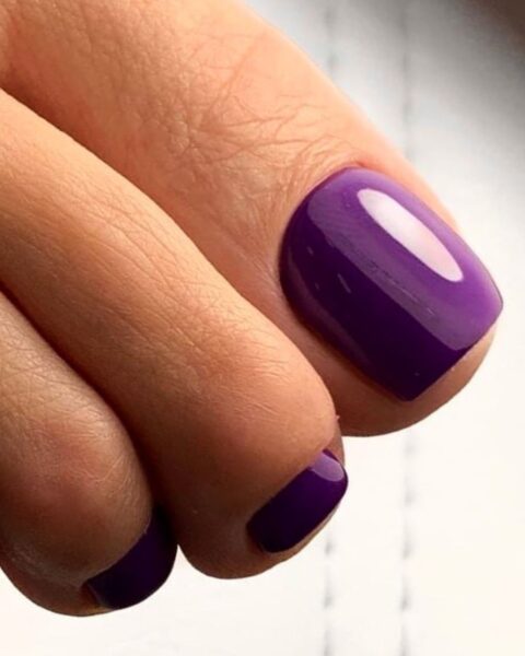 Intensywny fioletowy pedicure na zadbanych paznokciach u stóp. Paznokcie są idealnie opiłowane w kształt kwadratowy z zaokrąglonymi krawędziami i pokryte błyszczącym lakierem hybrydowym w żywym, purpurowym/elektrycznym odcieniu fioletu. Ujęcie z bliska, skupiające się na idealnym połysku i wykończeniu. Modny pedicure w kolorze 'Ultra Violet'.