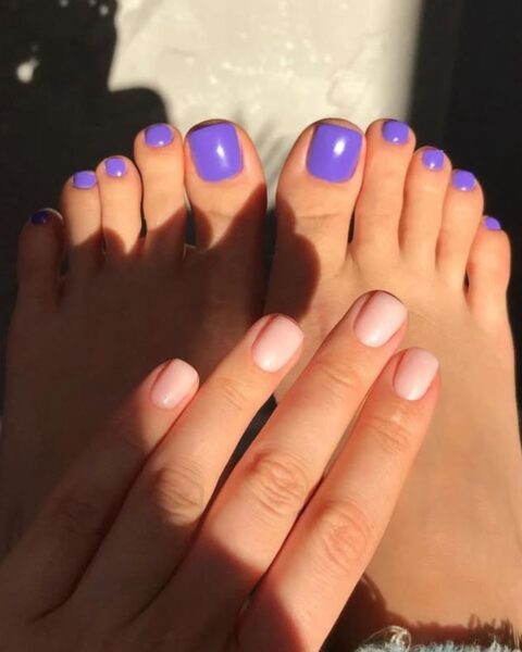 Letnie zestawienie manicure i pedicure. Paznokcie u stóp pomalowane na jasny, intensywny, lawendowy fiolet (lub liliowy). Dłoń widoczna na pierwszym planie ma manicure nude/baby boomer w delikatnym, cielistym różu. Całość w klimatycznym, kontrastowym świetle słonecznym, które rzuca cień stóp na ścianę. Osoba w jeansach z postrzępioną nogawką. Zadbana dłoń i stopy w modnych kolorach.
