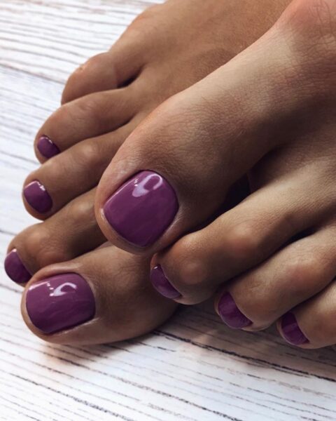 Pedicure hybrydowy w modnym, zgaszonym fiolecie z nutą różu (kolor "Violet Garden"). Paznokcie u stóp są starannie opracowane i pomalowane na jednolity, lśniący kolor. Ujęcie z bliska, prezentujące dbałość o detale i gładką skórę, na tle bielonej, drewnianej podłogi/tła. Kolor idealny do jesiennych stylizacji.