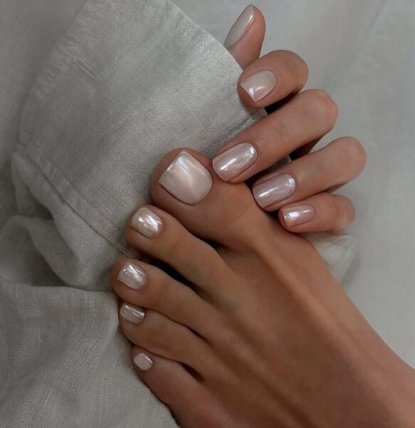 Harmonia manicure i pedicure w perłowym odcieniu nude z efektem „mirror glaze”. Paznokcie dłoni i stóp pomalowane tym samym lakierem w świetlistym, mlecznym kolorze z delikatnym opalizującym połyskiem. Tło z lnianej tkaniny dodaje naturalnego, subtelnego charakteru.