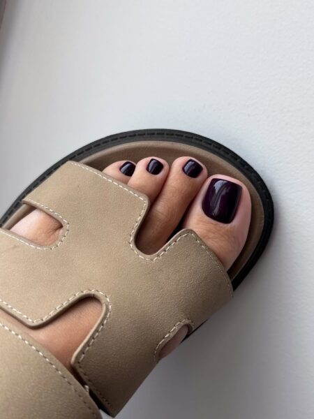 Pedicure na październik 2025: Stopa w beżowych, skórzanych sandałach typu slides. Paznokcie pomalowane intensywnym, głębokim odcieniem burgundowej czerni, które kontrastują z jasnym, neutralnym kolorem butów i minimalistycznym designem.