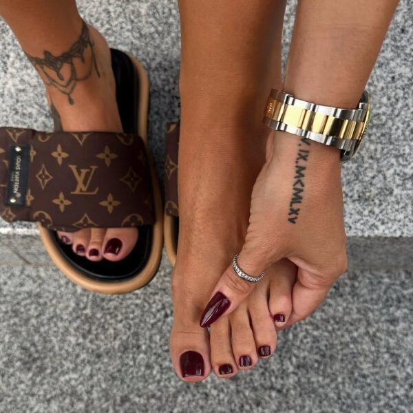 Dwie stopy w pedicure bordowym, sfotografowane na tle chodnika. Jedna stopa w luksusowych klapkach Louis Vuitton, druga przytrzymywana dłonią z dopasowanym manicure w tym samym odcieniu czerwieni. Widoczna także biżuteria – srebrny pierścionek oraz złoto-srebrny zegarek na nadgarstku, co podkreśla elegancję i spójność stylizacji.