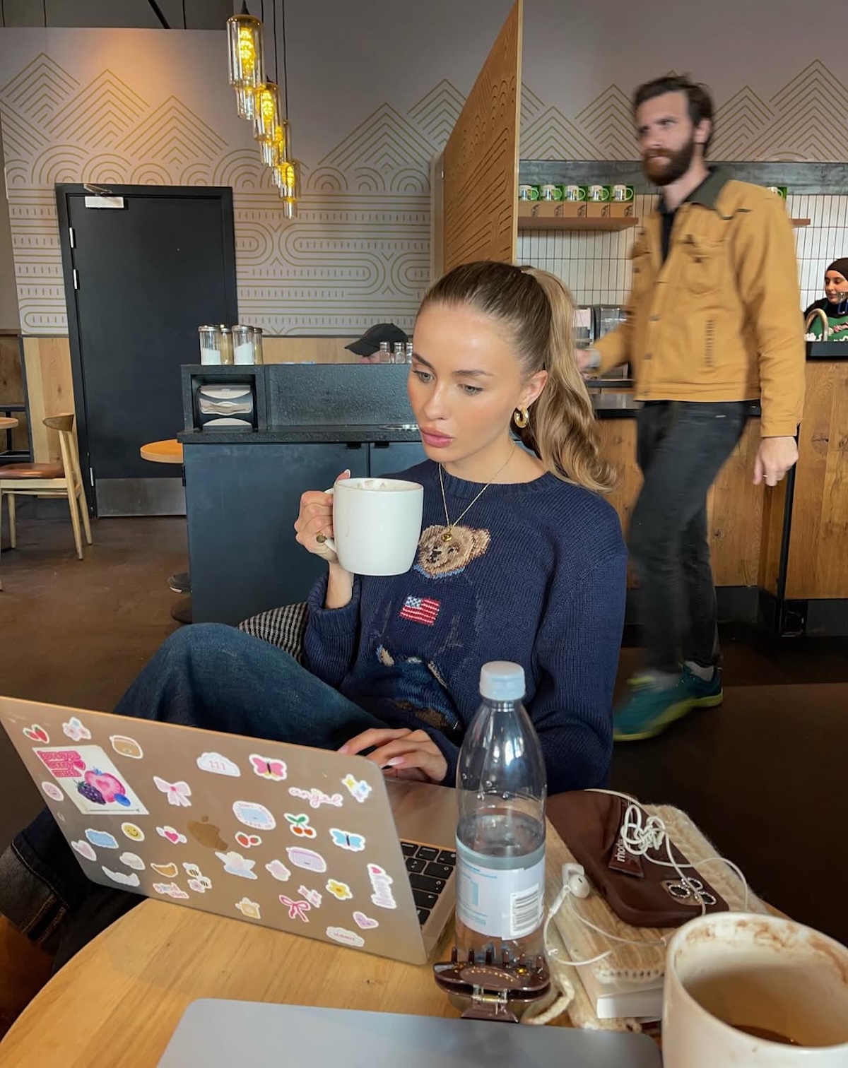 Netflix listopad 2025 seriale: Młoda kobieta siedząca w kawiarni przy drewnianym stoliku, z laptopem ozdobionym kolorowymi naklejkami i filiżanką kawy w dłoni. W tle nowoczesne wnętrze z industrialnym oświetleniem i geometrycznym wzorem na ścianie – atmosfera skupienia i codziennego rytuału pracy przy kawie.