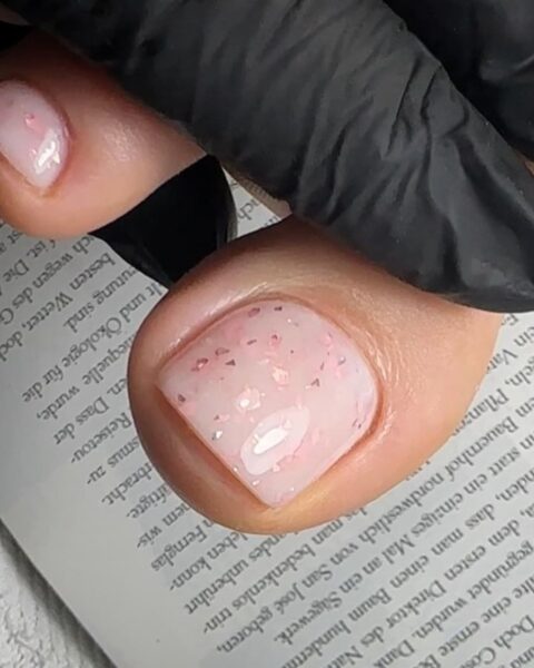 Pedicure w stylu jelly nails lub milk bath – transparentny, mleczno-różowy lakier z zatopionymi w środku drobinkami różowego brokatu (flakies). Ten subtelny, błyszczący efekt tworzy na paznokciach stóp delikatny, elegancki look i podkreśla dziewczęcą, świeżą estetykę.