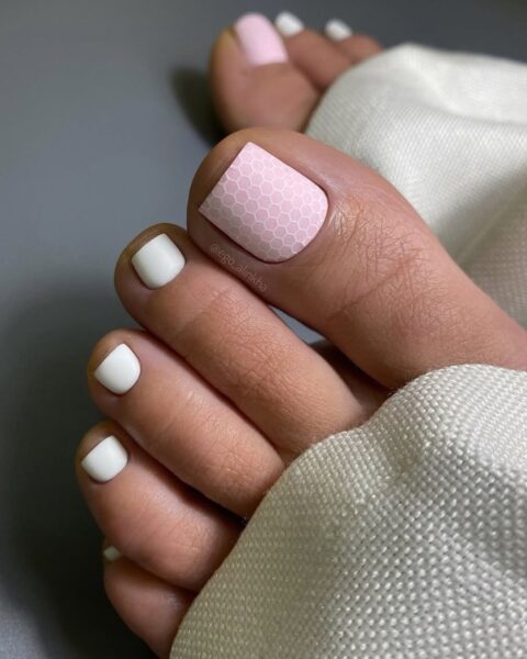 Nowoczesny pedicure łączący klasyczną biel z delikatnym, pudrowym różem na dużym palcu. Paznokieć ozdobiony jest modnym wzorem plastra miodu (honeycomb) lub geometrii, który nadaje subtelny, intrygujący akcent i wprowadza modową świeżość do klasycznej stylizacji.