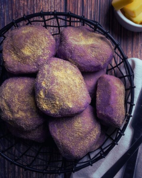 Zdjęcie w stylu rustykalnym, przedstawiające koszyk wypełniony fioletowymi bułeczkami ube pandesal, posypanymi żółtym pudrem. Obok, na lnianej serwetce, leżą czarny nóż i widelec. W tle widać drewniany blat i małą, białą miseczkę z kawałkami masła lub sera.