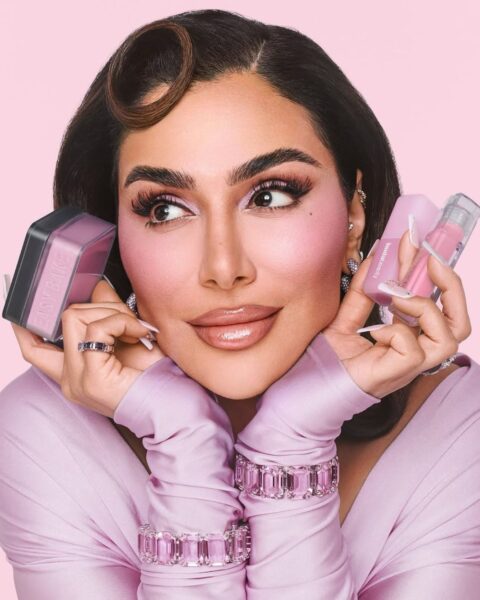 Influencerka modowa i makijażowa (Huda Kattan) pozująca na różowym tle. Kobieta ma na sobie pudroworóżową bluzkę oraz lśniącą, różową biżuterię. Prezentuje odcienie różowego makijażu z mocno zaakcentowanym różem na policzkach oraz trzyma w dłoniach dwa kosmetyki do makijażu.