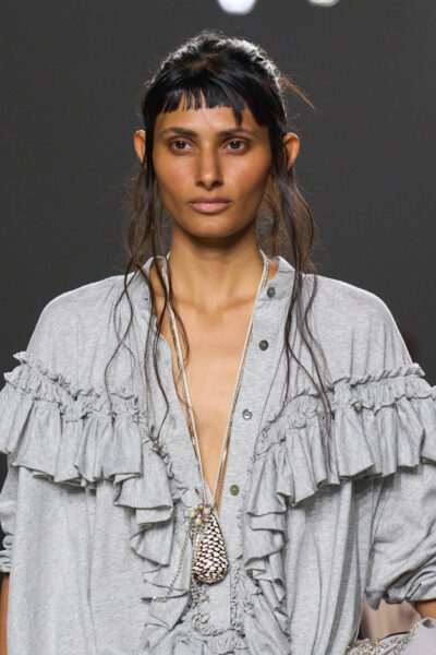 Zbliżenie na modelkę w szarej bluzie ozdobionej falbanami i metalicznym naszyjnikiem z motywem muszli. Połączenie casualowej miękkości z artystycznym detalem charakterystyczne dla eklektycznej estetyki Driesa Van Notena. Trendy wiosna 2026