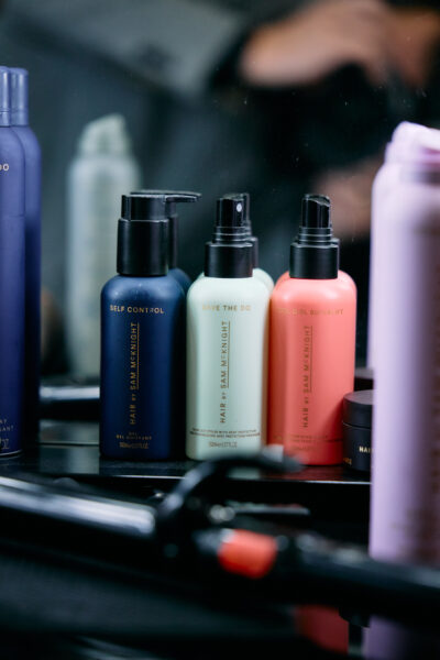 Zbliżenie na zestaw produktów do stylizacji włosów marki Hair by Sam McKnight ustawionych na toaletce w garderobie. Widać trzy butelki w odcieniach granatu, jasnej zieleni i różu z eleganckimi złotymi napisami. W tle odbicie w lustrze oraz sprzęt fryzjerski, tworzące atmosferę profesjonalnego backstage’u.
