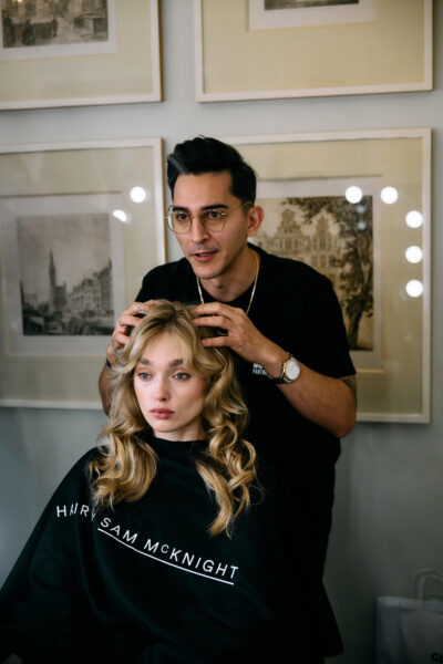 Stylista fryzur pracujący nad uczesaniem modelki o blond włosach w delikatnych falach, siedzącej w fotelu salonu z peleryną z napisem Hair by Sam McKnight. W tle klasyczne czarno-białe grafiki w ramach, a światło odbite od luster tworzy miękką, studyjną aurę.