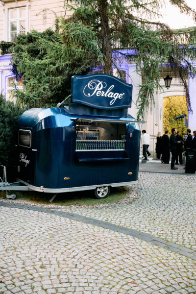 Stylowy niebieski food truck marki Perlage zaparkowany na dziedzińcu eleganckiej willi otoczonej zielenią. Wnętrze pojazdu wypełniają butelki wody mineralnej, a w tle goście w czarnych stylizacjach gromadzą się przy wejściu na wydarzenie.