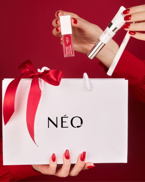 Zestaw NEO MAKE UP Classic X-Mas Look SET. Widok na dłonie chowające kosmetyki do białej torebki prezentowej z napisem NEO. Całość jest na czerwonym tle.