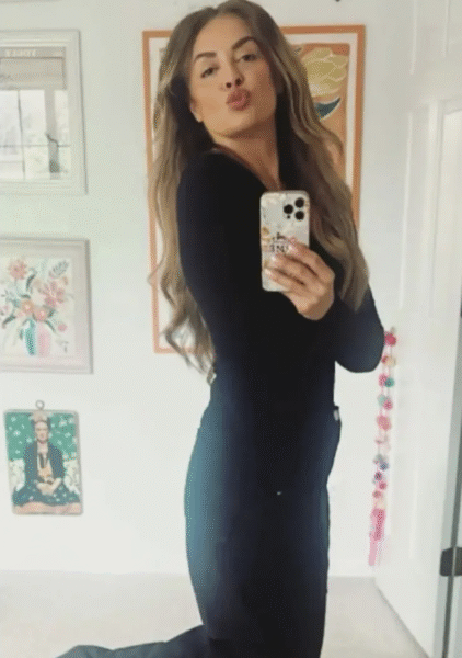 Kobieta robiąca selfie w lustrze, ubrana w dopasowany czarny kombinezon. Pozuje z uniesionymi ustami w geście pocałunku, w tle kolorowe dekoracje i ilustracje w domowym wnętrzu. Ujęcie pełne kobiecej energii i pewności siebie.
