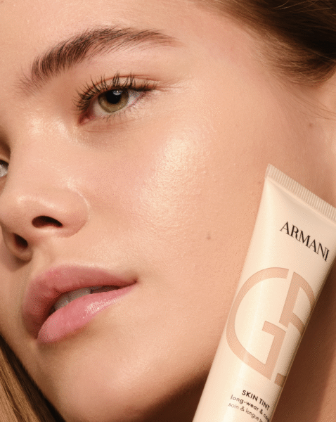 Zbliżenie twarzy modelki o naturalnym blasku, trzymającej beżową tubkę fluidu Armani Glow Skin Tint. Zdjęcie promuje efekt promiennej, zdrowej cery w duchu „no makeup” z luksusowym wykończeniem.