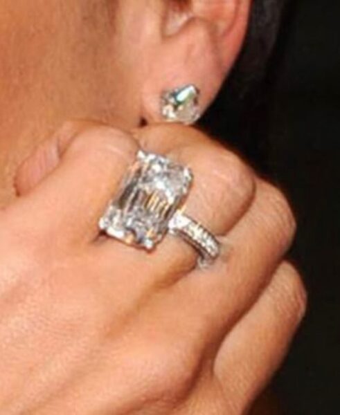 Zbliżenie na pierścionek zaręczynowy Victorii Beckham z ogromnym, prostokątnym diamentem w szlifie emerald, który połyskuje na tle diamentowych kolczyków.
