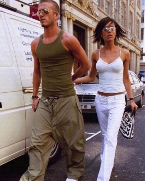 Victoria Beckham w białym topie na cienkich ramiączkach i białych spodniach podczas spaceru po Londynie z Davidem Beckhamem, który ma na sobie bojówki i sportową koszulkę, para trzyma się za ręce, prezentując styl z początku lat 2000.