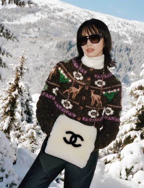 Modelka w zimowej scenerii Alp, ubrana w wełniany sweter z górskim motywem jeleni i choinek od Chanel, białą golfową bluzę oraz okulary przeciwsłoneczne oversize. W dłoniach trzyma futrzaną torebkę z czarnym logo CC na tle śnieżnych szczytów.