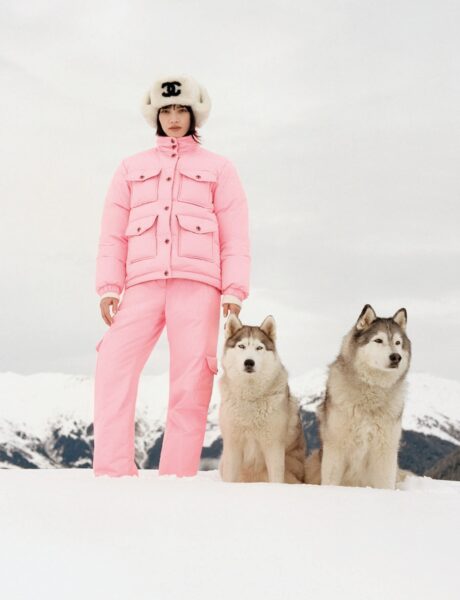 Modelka w różowym kombinezonie narciarskim Chanel i białej futrzanej czapce z logo CC pozuje na śniegu obok dwóch psów husky. Minimalistyczna sceneria i pastelowy róż kontrastują z surowym pejzażem gór.