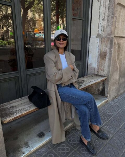 Cigarette jeans na jesień 2025. Kobieta siedzi na drewnianej ławce na zewnątrz, ubrana w oversize'owy beżowy płaszcz (lub trencz), biały T-shirt, ciemne dżinsy z postrzępionym dołem i czarne mokasyny. Stylizację uzupełnia sportowa czapka z daszkiem i okulary przeciwsłoneczne. Obok niej leży czarna zamszowa torebka.