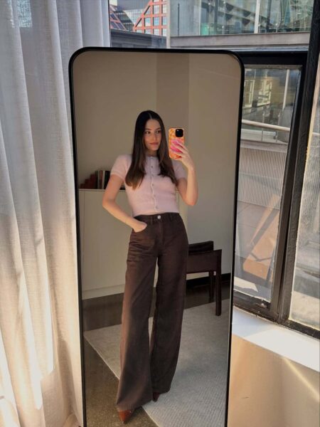 Jasnobrązowe lub czekoladowe jeansy na jesień 2025 z szerokimi nogawkami, zestawione z krótkim, jasnoróżowym, zapinanym sweterkiem. Kobieta stoi przodem do lustra w pomieszczeniu, prawdopodobnie salonie lub sypialni, z widokiem na miasto przez duże okno. Jej długie, ciemne włosy są rozpuszczone i opadają na ramiona. Stylizacja jest nowoczesna i casualowa, idealna na przejściową pogodę.