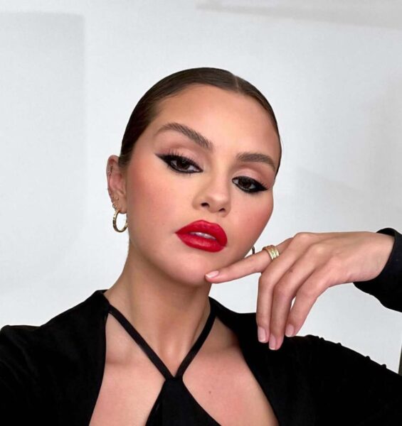 Portret Seleny Gomez z gładko zaczesanymi włosami i odważnym makijażem. Ma wyraźne, graficzne czarne kreski na powiekach i głęboką czerwień na ustach, co podkreśla jej styl. Na palcu nosi duży, złoty pierścień, a w uszach złote kolczyki koła. Aktorka pozuje, podpierając brodę dłonią. Intensywny kolor czerwonej szminki stanowi centralny punkt tego eleganckiego looku.