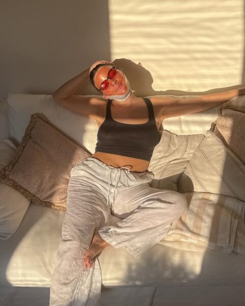 Zdjęcie kobiety relaksującej się na jasnej kanapie w promieniach zachodzącego słońca. Ma na sobie czarny top na ramiączkach i luźne, jasnoszare spodnie. Na twarzy ma nałożoną czerwoną maseczkę LED lub kolorowe okulary przeciwsłoneczne i biały bandaż lub element pielęgnacyjny owinięty wokół brody, co jest popularnym motywem w pielęgnacji anti-aging. Kobieta ma związane włosy i opiera się o poduszki, ciesząc się złotą godziną. Ta poza i pielęgnacja nawiązują do trendów self-care i urodowych trików z tiktoka na domowe SPA i błyskawiczną regenerację.