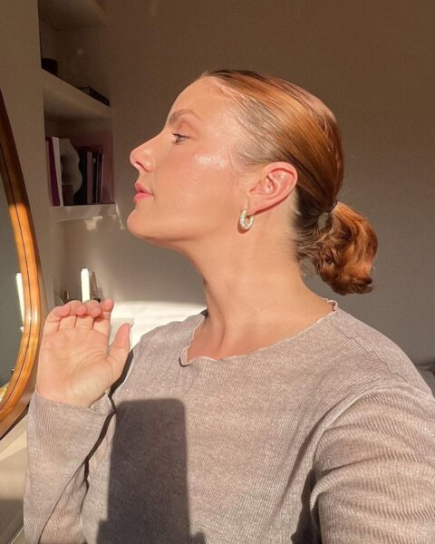 Ujęcie profilowe kobiety o rudawych włosach, zrobione w promieniach zachodzącego słońca, które podkreśla jej lśniącą, zdrową skórę. Włosy są zebrane w niski kucyk, z gładko zaczesanymi pasmami w stylu "sleek look". Kobieta ma na sobie beżowy, prążkowany sweter i złotą kolczyki-koła. Podniesiona pozycja głowy i dłoni skierowana ku światłu akcentuje nawilżoną, pełną blasku cerę, co jest efektem popularnych urodowych trików z tiktoka na "glass skin" i efekt rozświetlenia przy użyciu balsamu lub olejku.