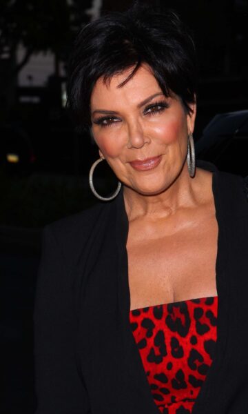 Zdjęcie z bliska przedstawia Kris Jenner uśmiechającą się lekko, z jej charakterystycznymi, krótkimi, ciemnymi włosami. Ma na sobie czarną marynarkę lub żakiet i top w czerwono-czarny wzór panterki. Nosi duże, srebrne kolczyki w kształcie kół oraz ma nałożony mocny makijaż oczu. Tło jest ciemne i niewyraźne, sugerując, że zdjęcie zostało zrobione w nocy lub w słabym oświetleniu.