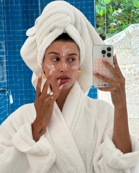 Selfie w łazience, przedstawiające Hailey Bieber w białym, frotowym szlafroku i białym ręczniku na głowie. Kobieta ma na sobie duże, złote kolczyki i duży pierścionek z brylantem. Nakłada palcami biały krem lub maseczkę na twarz, na której widać już kilka kropek produktu. Tło stanowią intensywnie niebieskie, błyszczące kafelki. Jest to scena z rutyny pielęgnacyjnej, która prawdopodobnie wykorzystuje produkty bogate w składniki aktywne, zgodnie z kompleksowym przewodnikiem po składnikach aktywnych dla zdrowej cery.