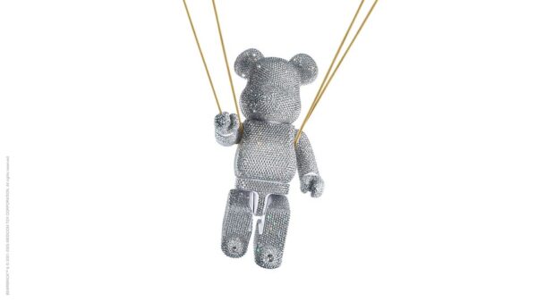 Artystyczny wisiorek w kształcie misia, cały pokryty kryształkami, zawieszony na złotych sznurkach. Obiekt przypomina designerską biżuterię lub figurkę kolekcjonerską, łącząc zabawny motyw z luksusową estetyką.