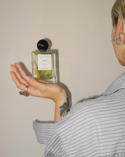 Ujęcie z tyłu przedstawiające kobietę z krótko ściętymi, jasnymi włosami i kolczykami-kółkami, ubraną w koszulę w paski, trzymającą w dłoni kwadratowy flakon perfum. Płyn wewnątrz flakonu ma zielonkawo-żółty kolor, a na etykiecie widnieje nazwa "VIOLA". Na ręce widoczny jest tatuaż i srebrny pierścień. Tło jest jednolite, jasne i neutralne.