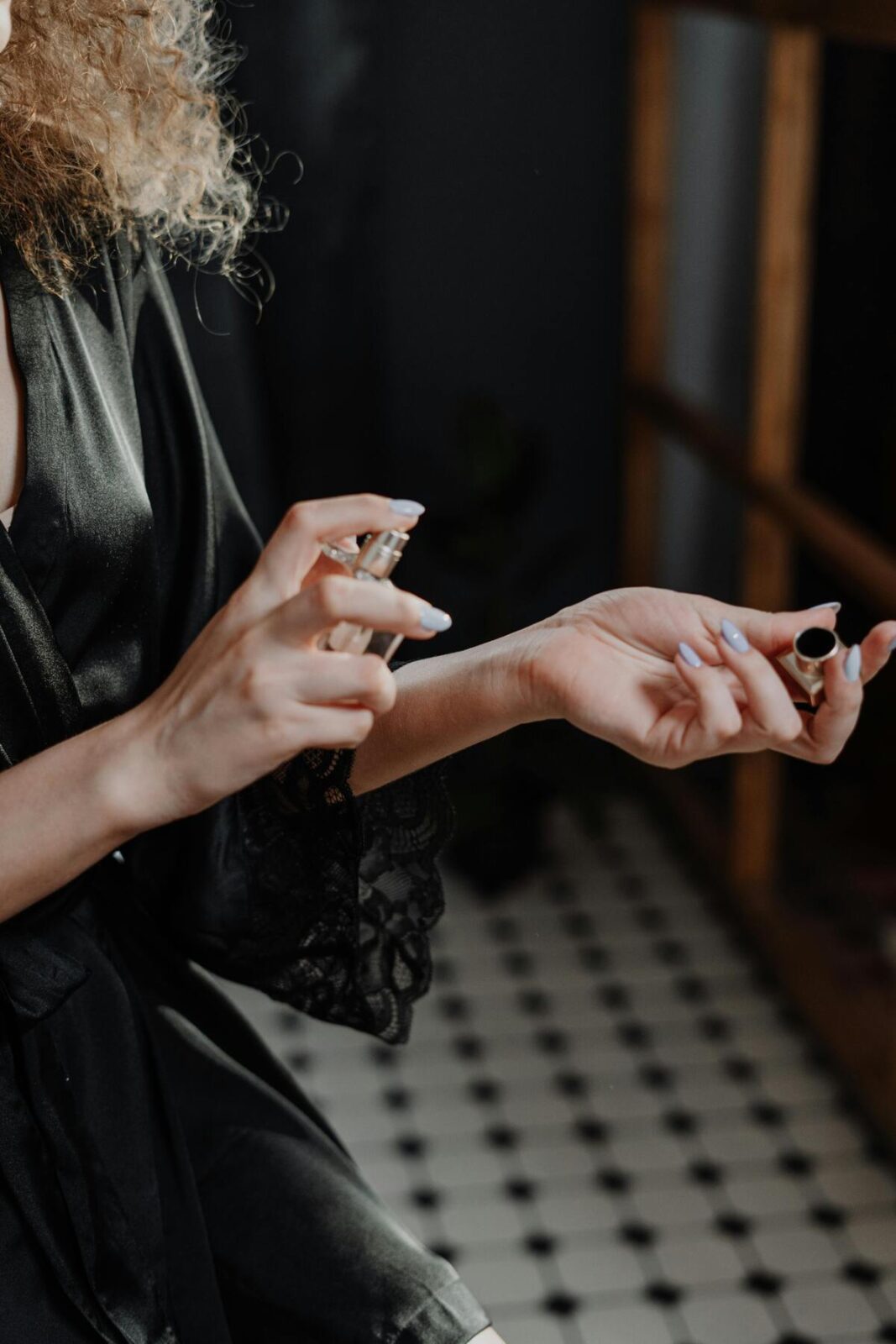 Jak świadomie wybierać perfumy? Trendy zapachowe 2025. Zbliżenie na dłonie kobiety w czarnym, satynowym szlafroku z koronkowym wykończeniem, która spryskuje perfumami wewnętrzną stronę nadgarstka, wykonując rytuał świadomego wyboru perfum. Ten gest symbolizuje znaczenie aplikacji zapachu w trendach zapachowych 2025. W tle widoczna jest czarno-biała, geometryczna podłoga i częściowo rozmyte, naturalne światło.