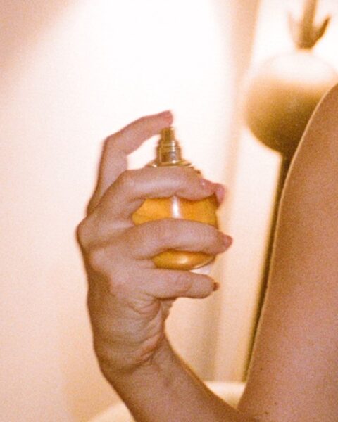 Jak świadomie wybierać perfumy? Zbliżenie na dłoń trzymającą mały flakon perfum ze złotym płynem i atomizerem, co symbolizuje świadomy wybór perfum i rytuał ich aplikacji. Ciepłe, bursztynowe kolory i miękkie, nastrojowe oświetlenie sugerują intymność związaną z wyborem osobistego zapachu, który może wpisywać się w ciepłe i otulające trendy zapachowe 2025. Zdjęcie ma ziarnisty, retro charakter.