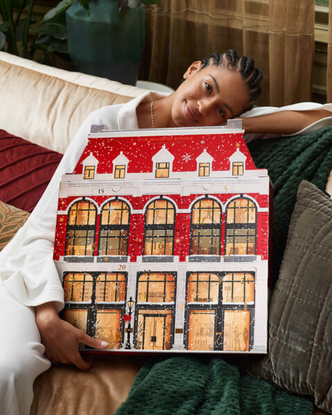 Uśmiechnięta kobieta siedzi na sofie i przytula duży, czerwono-biały kalendarz adwentowy w kształcie kamienicy. Ma na sobie białą koszulę i warkoczyki. Tło wypełniają poduszki i zielony koc.
