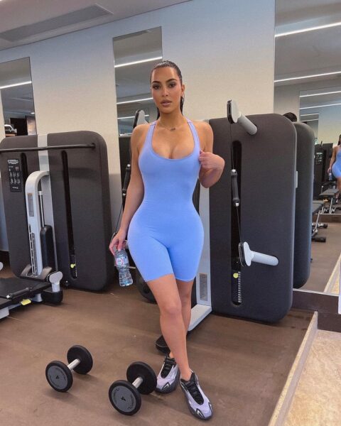 Kim Kardashian pozuje w siłowni, stojąc przed maszynami treningowymi i hantlami. Ma na sobie obcisły, jasnoniebieski kombinezon sportowy i sportowe buty. Trzyma w ręce butelkę wody.