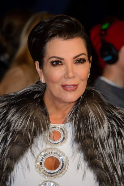 Zdjęcie z bliska przedstawia Kris Jenner ubraną w biały strój, na który narzucony jest luksusowy, duży kołnierz lub futrzana etola w kolorach srebrno-czarnym (lub szarym). Kołnierz jest puszysty i otacza jej szyję. Na białej sukience widać duże, okrągłe aplikacje lub naszyjniki wysadzane błyszczącymi kryształami. Kris ma ciemne, elegancko zaczesane włosy, mocny makijaż oczu i drobne kolczyki w uszach. Uśmiecha się lekko. W prawym górnym rogu widać niewyraźny fragment postaci w czerwonej czapce i słuchawkach.
